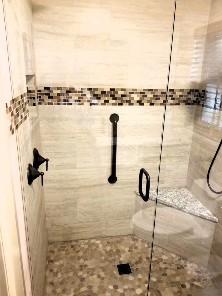 Walk-in shower with clear glass doors, grab bars, corner seat, and pebble floor tile
